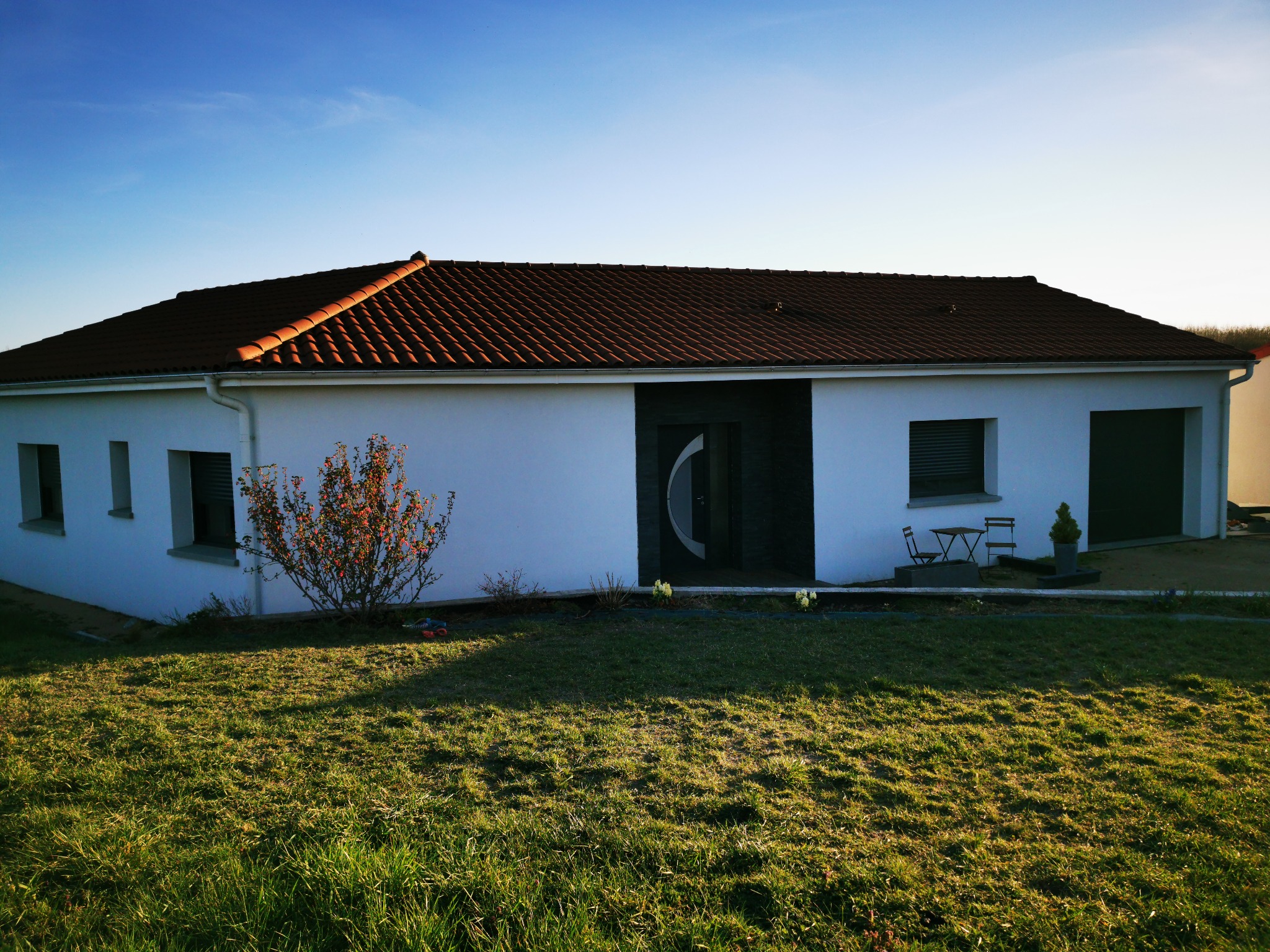 Parterowy, biały dom z brązowym dachem w słoneczny dzień. Widoczne okna, drzwi z elementami szkła, trawnik i niewielki ogródek przed domem. Minimalistyczny styl.