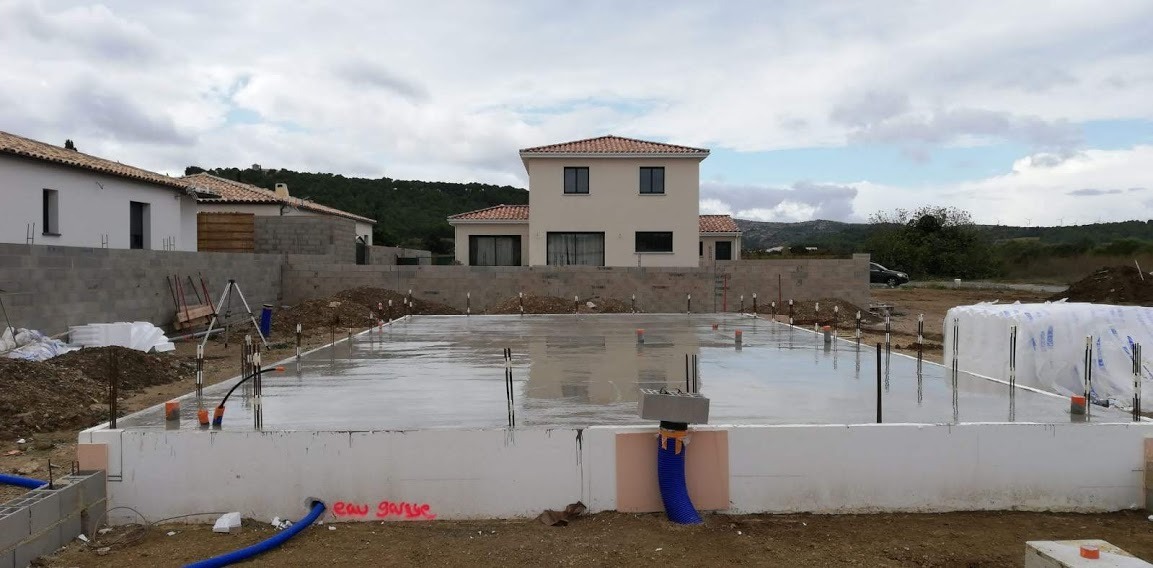 Wylana płyta fundamentowa pod budowę domu jednorodzinnego. Widoczne zbrojenia, instalacje i bloczki fundamentowe. W tle budynki w trakcie budowy i krajobraz.