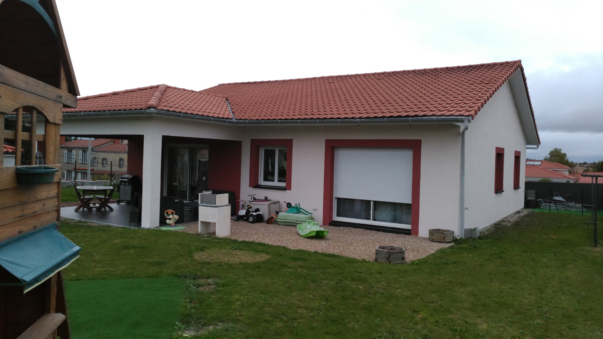 Parterowy dom z czerwonym dachem i obramowaniami okien. Na posesji trawnik i zabawki dziecięce. Widoczny fragment drewnianego placu zabaw.