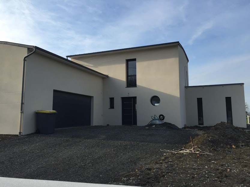 Nowoczesny dom z garażem i okrągłym oknem, widok z zewnątrz. Na podjeździe żwir i taczka. Minimalistyczna architektura, jasna elewacja.