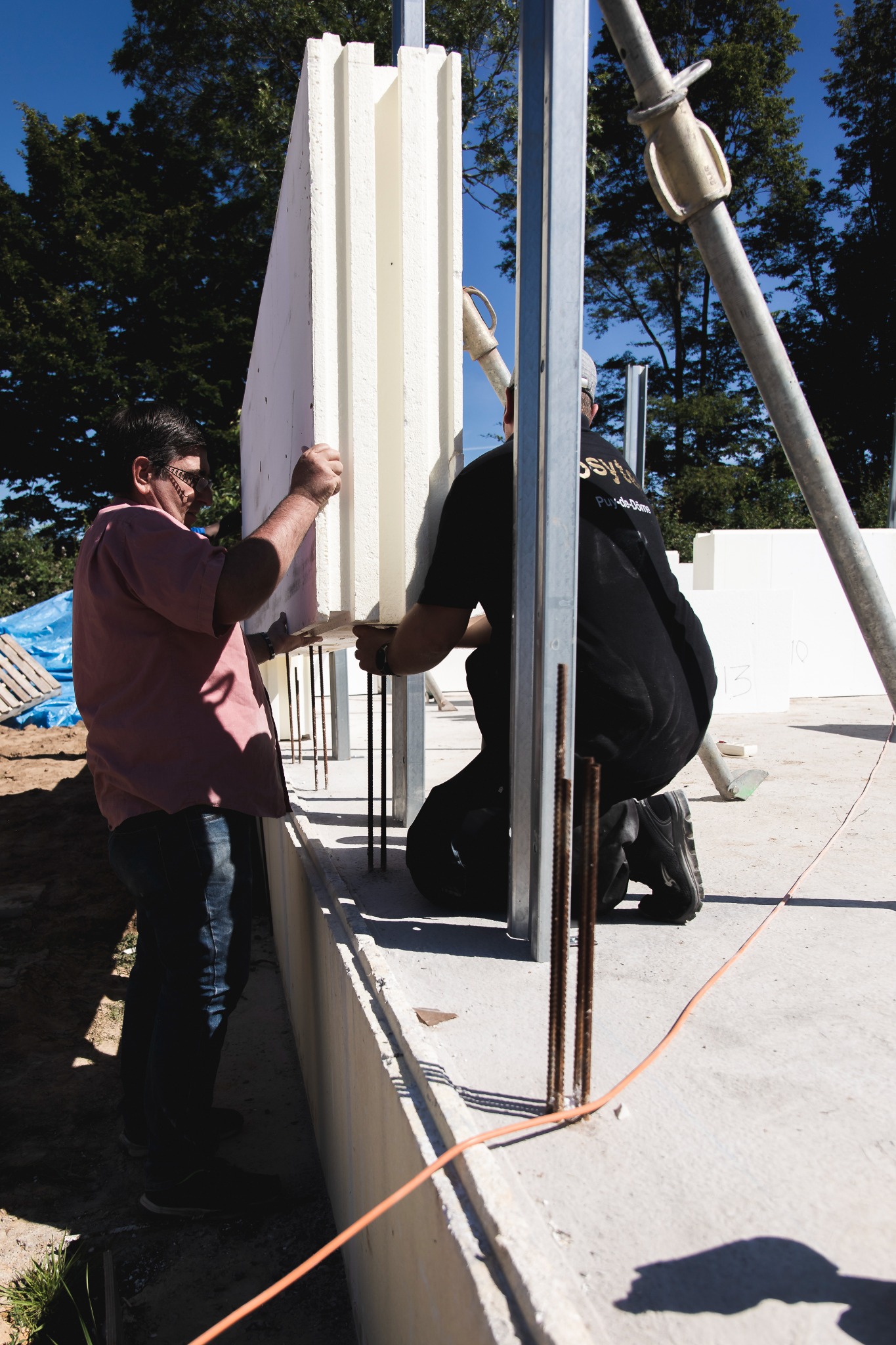 Montaż białych paneli izolacyjnych na fundamencie z wystającymi prętami zbrojeniowymi. Dwóch robotników przy pracy, widok z bliska.