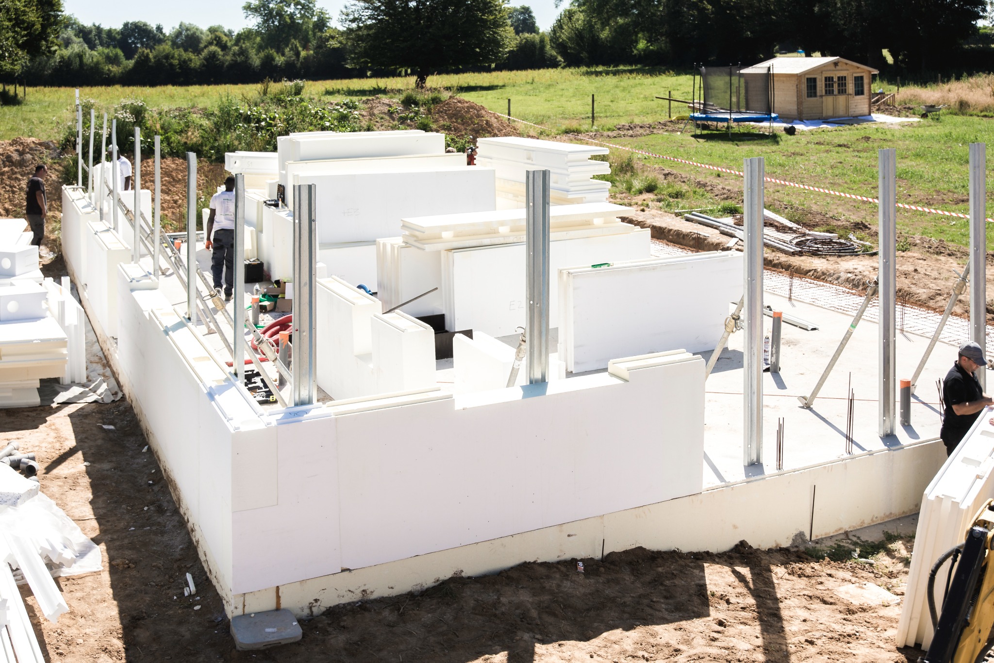 Budowa domu z białych paneli izolacyjnych, widoczne metalowe podpory i pracownicy. W tle zielone pole i mały drewniany budynek.