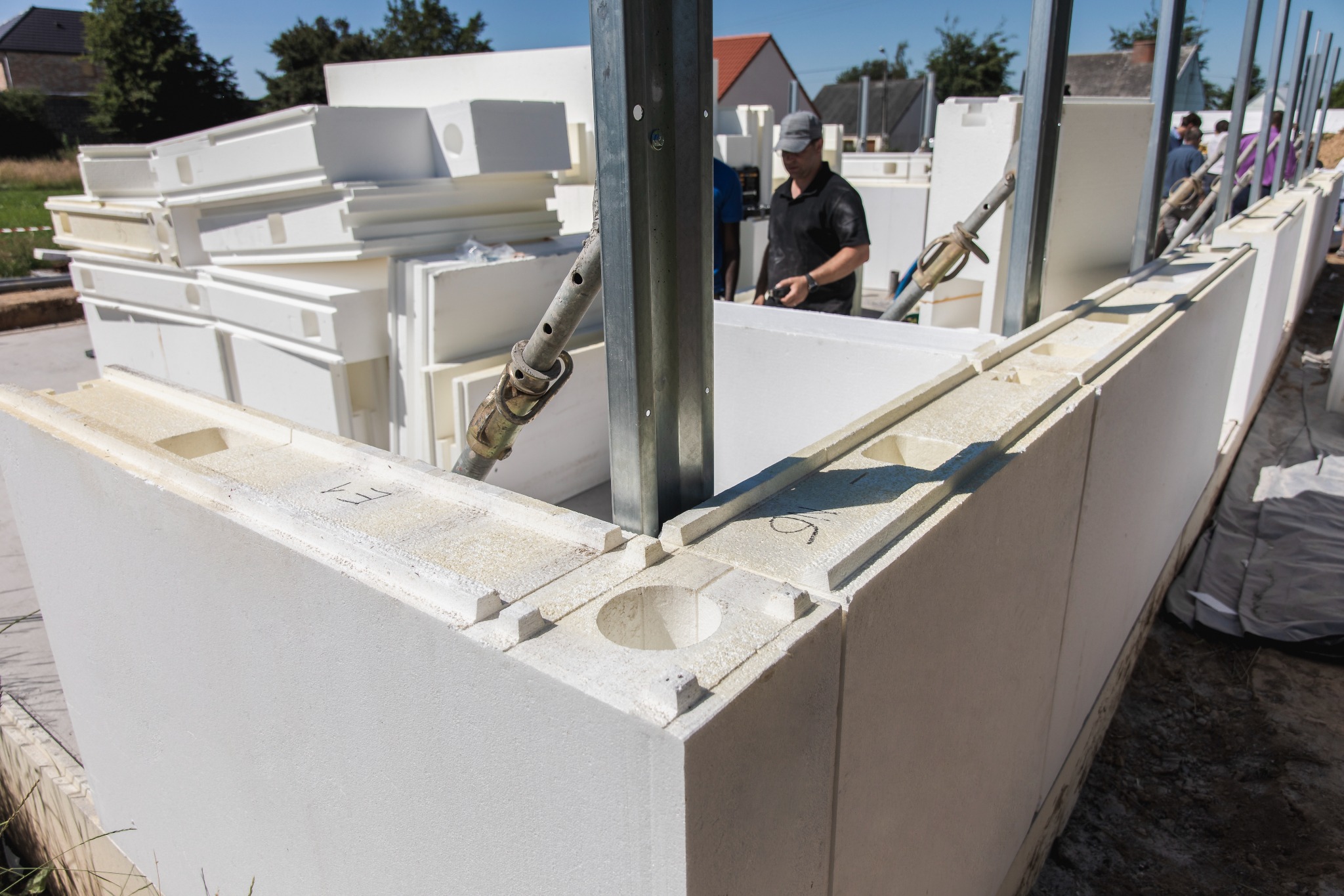 Wznoszenie ściany z białych bloczków styropianowych, z widocznymi otworami, wspartych metalowymi stemplami. W tle pracownik w czarnej koszulce i czapce.