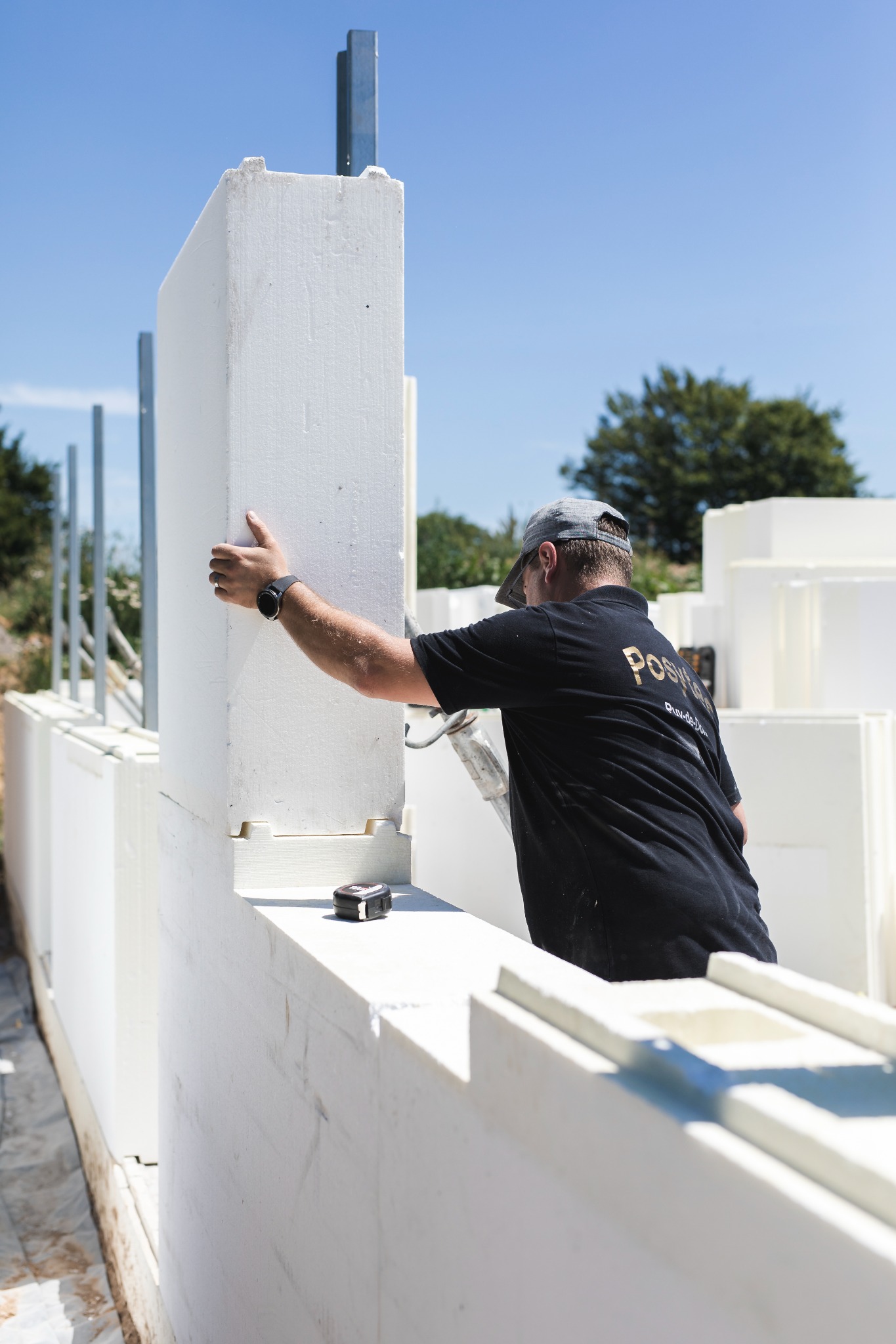 Montaż białych paneli ściennych z pianki przez pracownika w czarnej koszulce na budowie domu modułowego w słoneczny dzień.