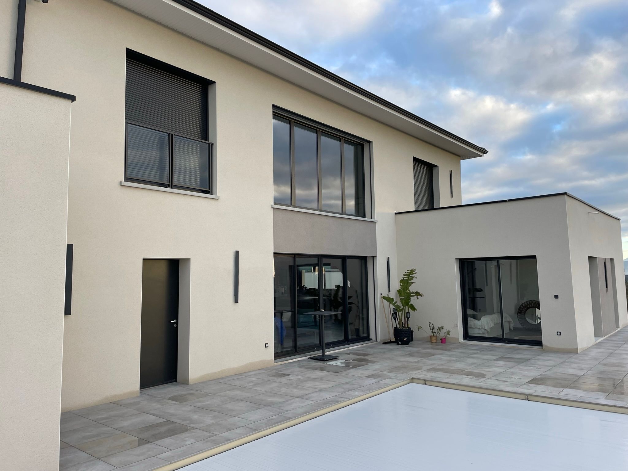 Nowoczesny dom z płaskim dachem, jasną elewacją i dużymi oknami. Widoczny taras z szarą kostką i przykryty basen. Minimalistyczny design i spokojna kolorystyka.