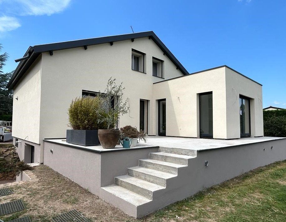 Jasny dom z dobudówką i schodami na taras. Uwagę przykuwają duże okna i minimalistyczny styl. Zieleń w donicach dodaje naturalnego akcentu.