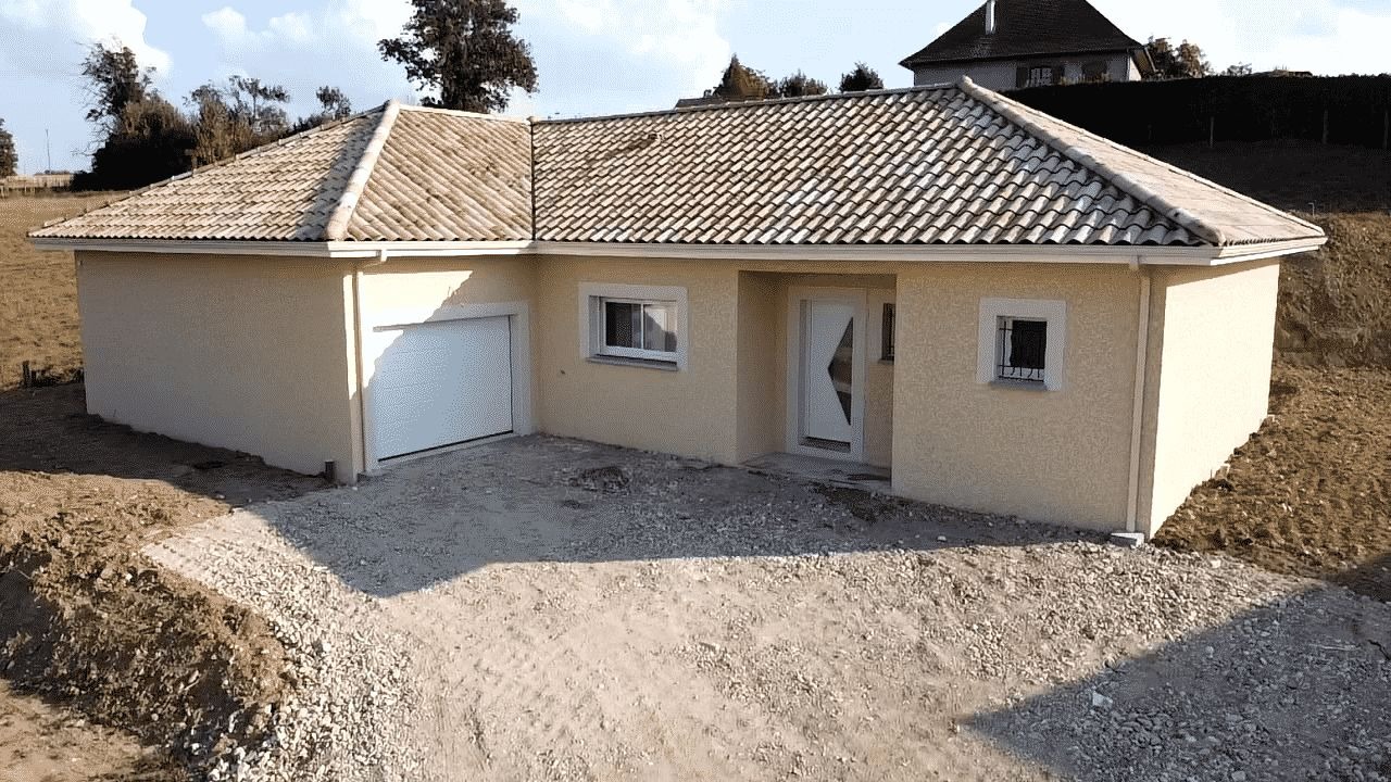 Parterowy dom z beżową elewacją i garażem, dach pokryty dachówką ceramiczną. Teren wokół domu przygotowany pod zagospodarowanie.