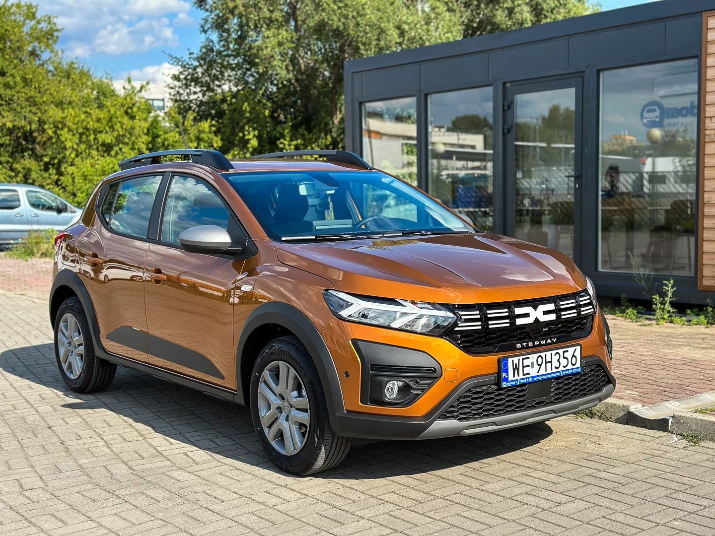 Pomarańczowa Dacia Sandero Stepway na kostce brukowej, z widoczną tablicą rejestracyjną i budynkiem w tle, prezentowana jako oferta wypożyczalni.