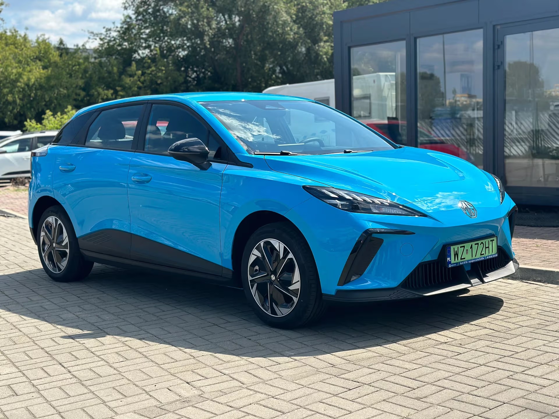 Błękitny, elektryczny MG 4 na tle przeszklonego budynku, z widoczną tablicą rejestracyjną. Auto stoi na brukowanym placu w słoneczny dzień.