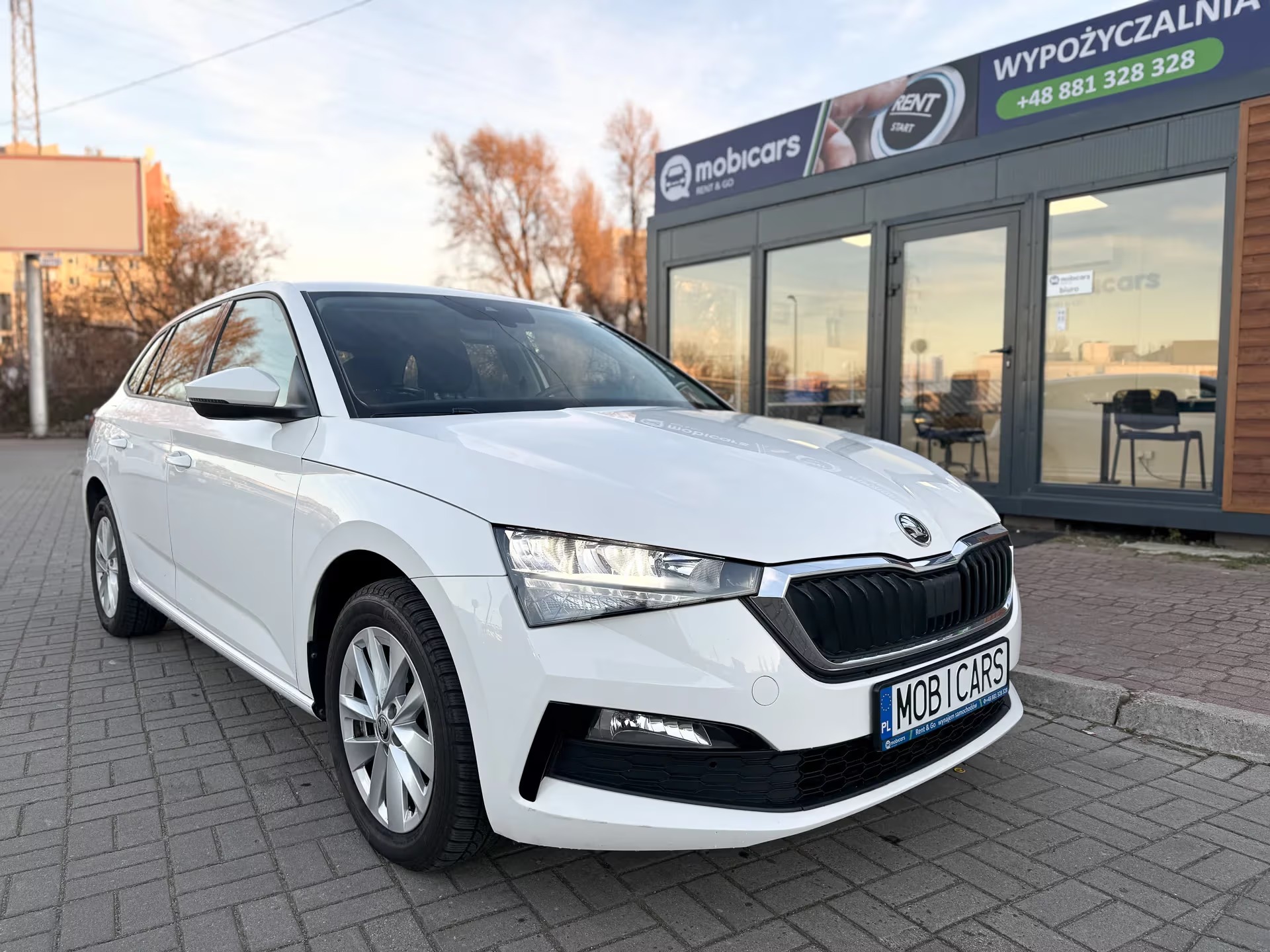 Biała Skoda Scala przed wypożyczalnią MobiCars. Widoczny numer telefonu. Auto na brukowanym chodniku, w tle budynek wypożyczalni i drzewa.