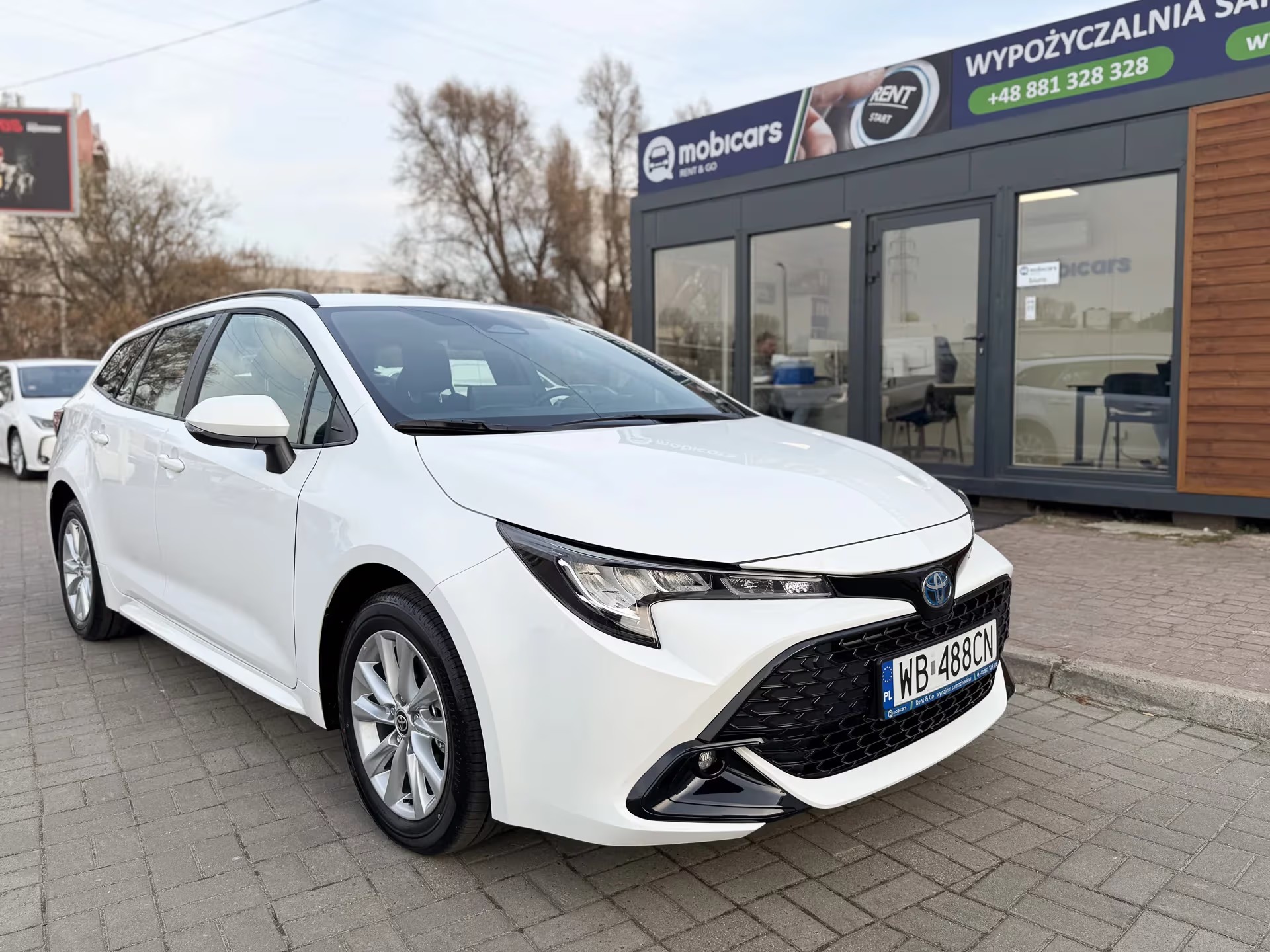 Biała Toyota Corolla kombi na tle biura wypożyczalni MobiCars. Widoczny numer rejestracyjny WB 488CN i logo firmy.