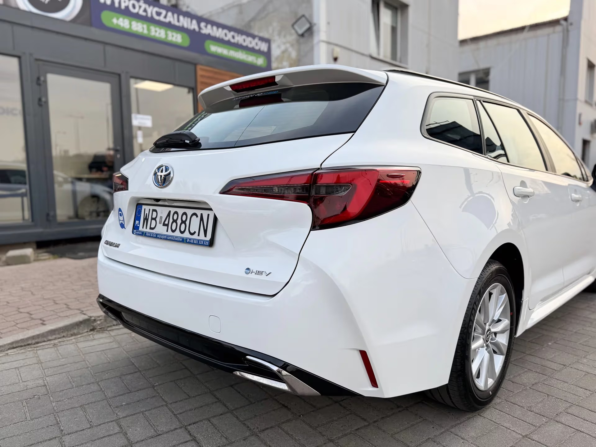 Biała Toyota Corolla kombi z widocznym numerem rejestracyjnym WB 488CN, zaparkowana przed budynkiem wypożyczalni samochodów, widok z tyłu, lekki kąt.