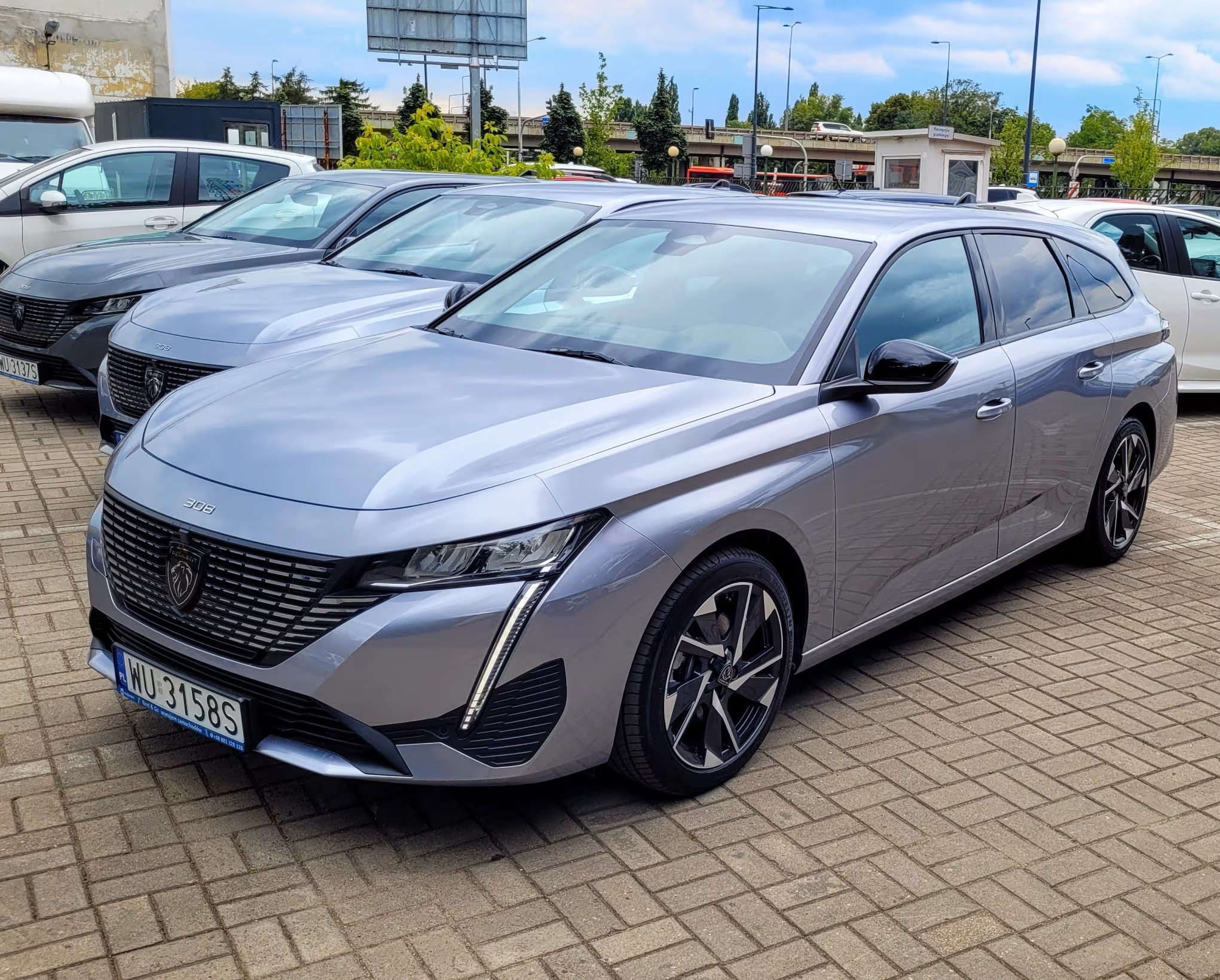 Szary Peugeot 308 SW na brukowanym parkingu. Widoczne kolejne auta w tle, budka parkingowa i fragment wiaduktu. Charakterystyczny grill i reflektory.