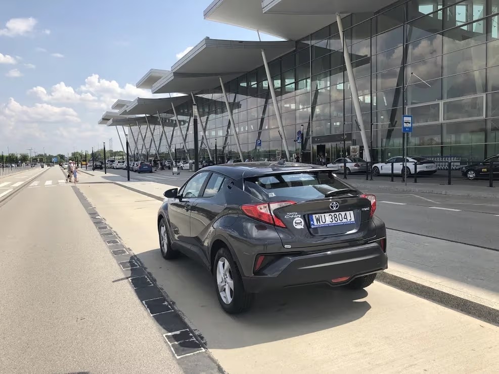Grafitowa Toyota C-HR zaparkowana przed nowoczesnym budynkiem lotniska, widok z tyłu, tablica rejestracyjna WU 3804J, jasne światło słoneczne.