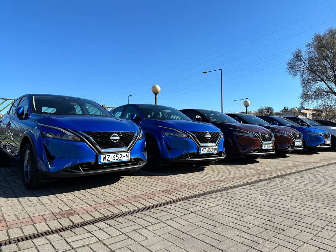 Rząd aut Nissan Qashqai w kolorach niebieskim i bordowym, zaparkowanych na kostce brukowej, z widocznymi numerami rejestracyjnymi i błękitnym niebem w tle.