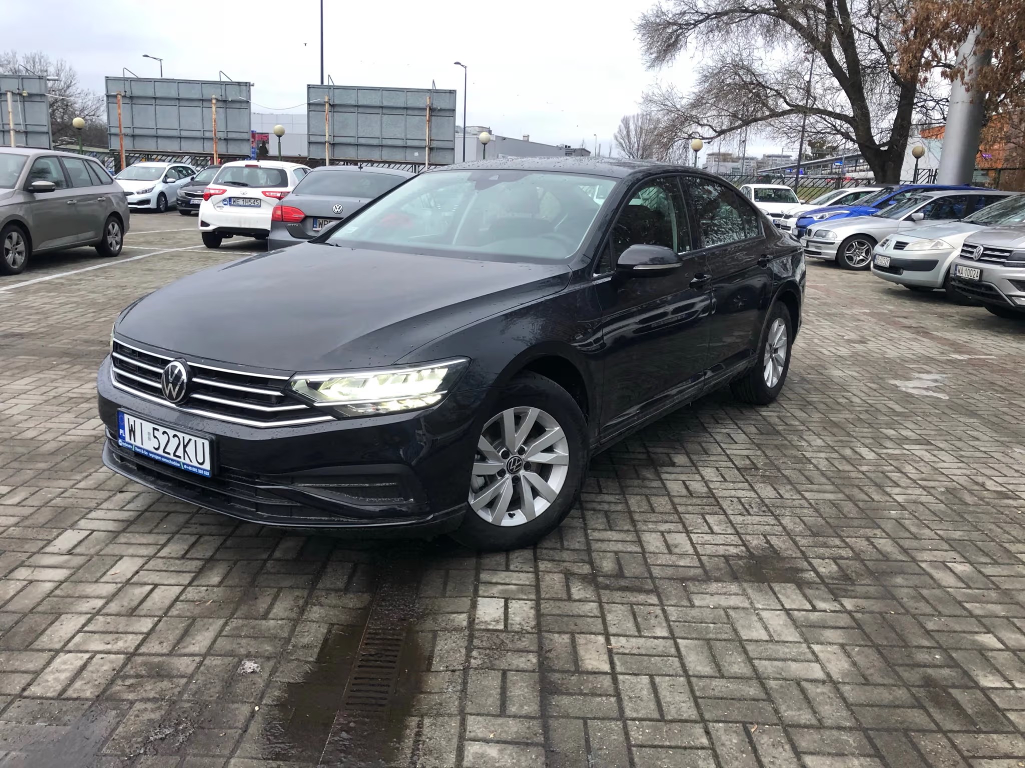 Czarny Volkswagen Passat na parkingu, oświetlony światłami dziennymi. Widoczne inne zaparkowane auta i kostka brukowa. Zdjęcie wykonane w pochmurny dzień.
