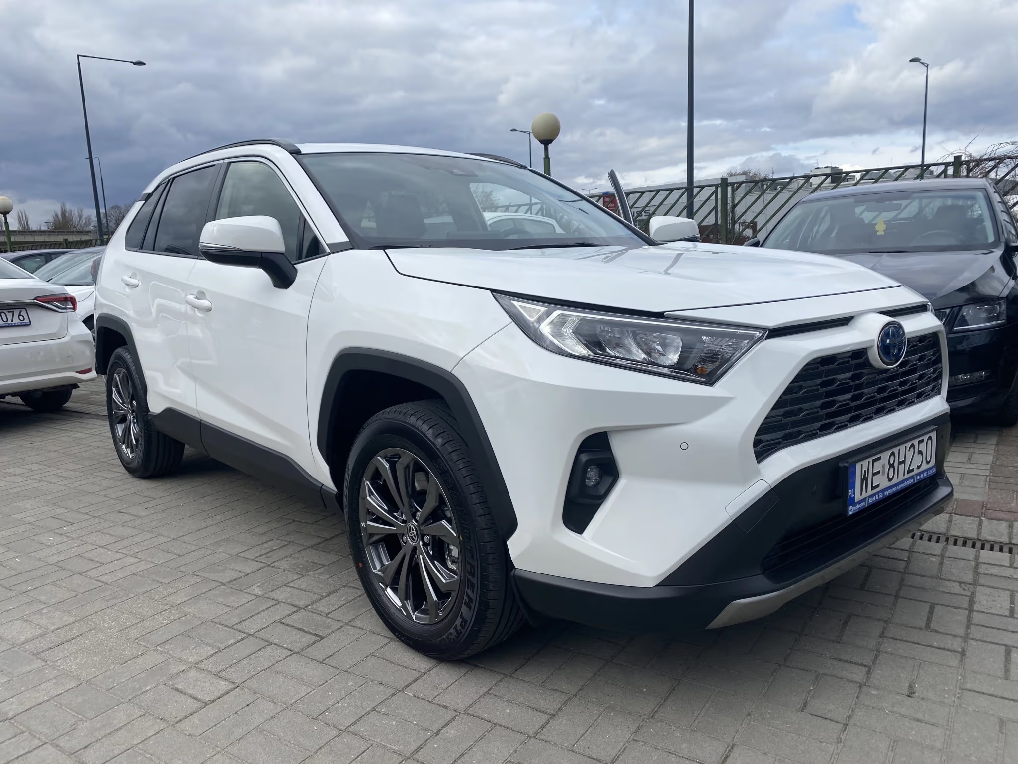 Biała Toyota RAV4 Hybrid na brukowanym parkingu, z otwartymi drzwiami kierowcy, widoczną tablicą rejestracyjną i fragmentami innych aut. Pochmurne niebo w tle.