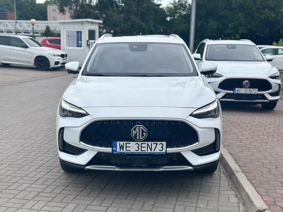 Biały MG HS, widok z przodu na tle kostki brukowej i innych aut. Tablica rejestracyjna widoczna. Auto na wynajem bez kaucji.