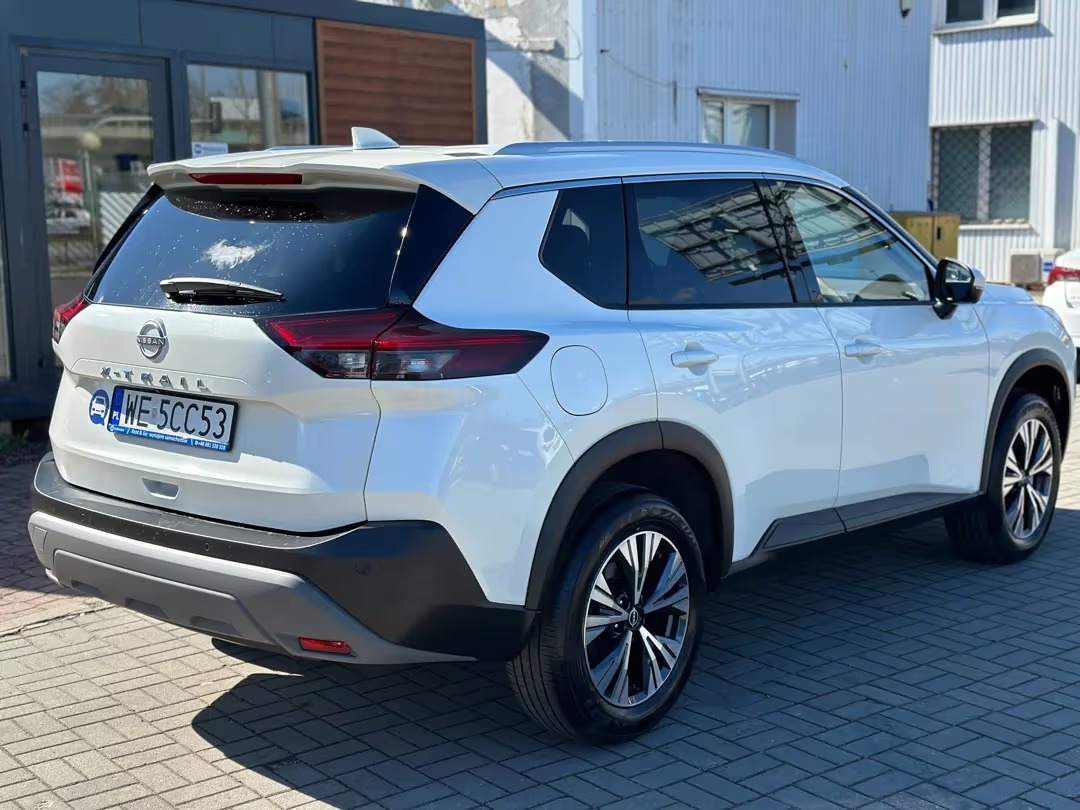 Biały Nissan X-Trail, 7-osobowy SUV z widocznym tyłem i tablicą rejestracyjną. Auto zaparkowane na kostce brukowej, jasne oświetlenie.