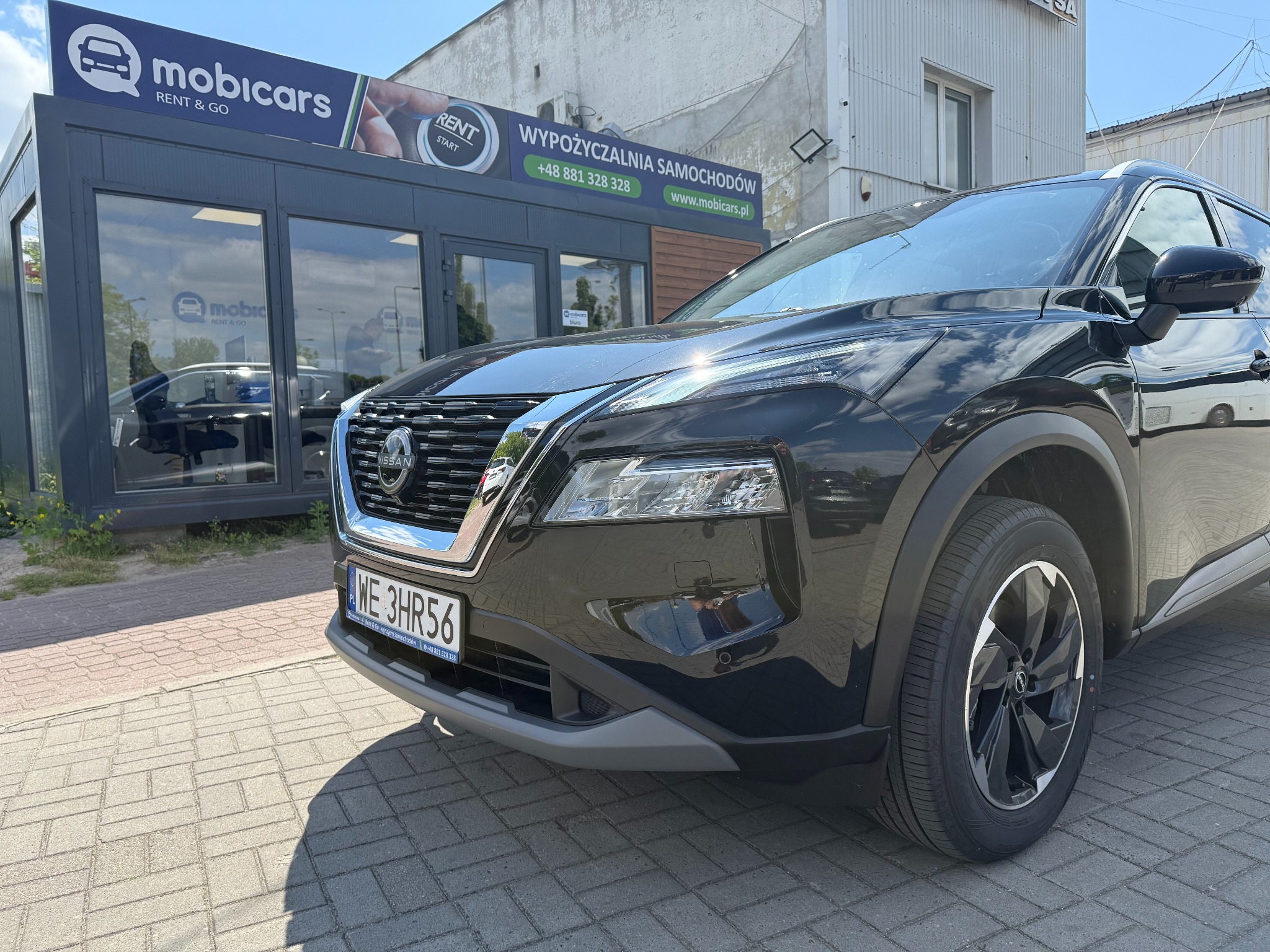 Czarny Nissan X-Trail przed biurem MobiCars, z widocznym numerem rejestracyjnym i oznaczeniami wypożyczalni. Ujęcie z bliska, podkreślające detale auta.