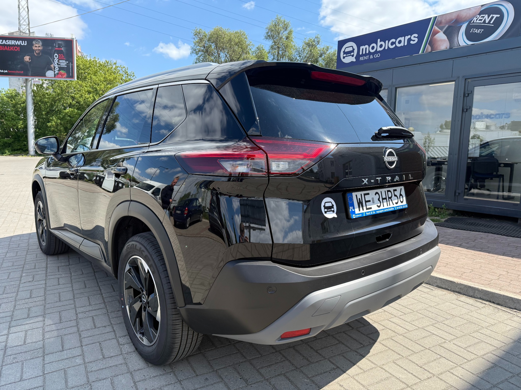 Czarny Nissan X-Trail, tył pojazdu, na tle biura MobiCars w Warszawie. Widoczny numer rejestracyjny, logo Nissan i oznaczenie modelu. Auto na wynajem bez kaucji.