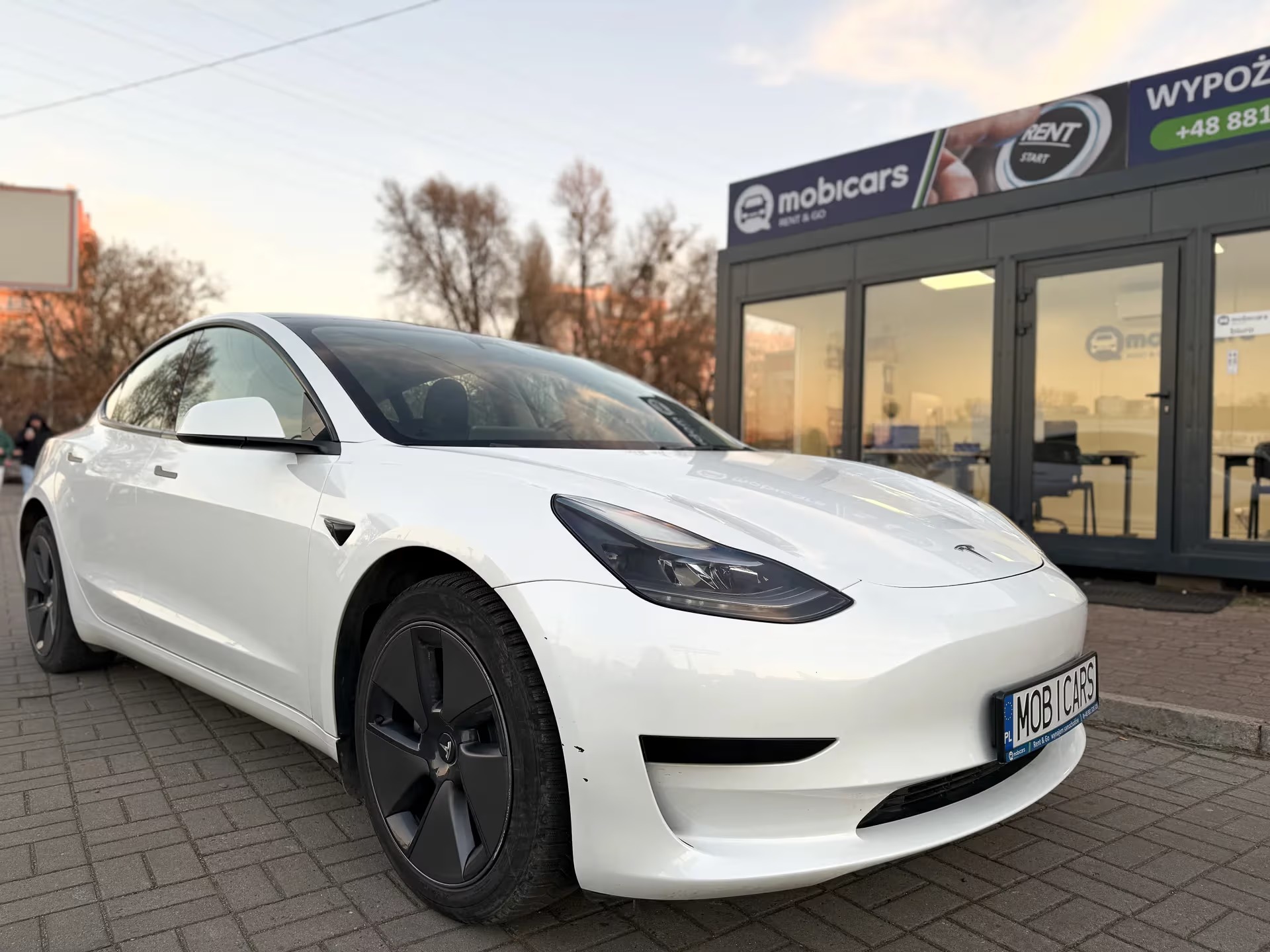 Biała Tesla Model 3 przed biurem MobiCars z szyldem 'Wynajem'. Elektryczny samochód na tle kostki brukowej i jesiennych drzew. Tablica rejestracyjna z nazwą firmy.
