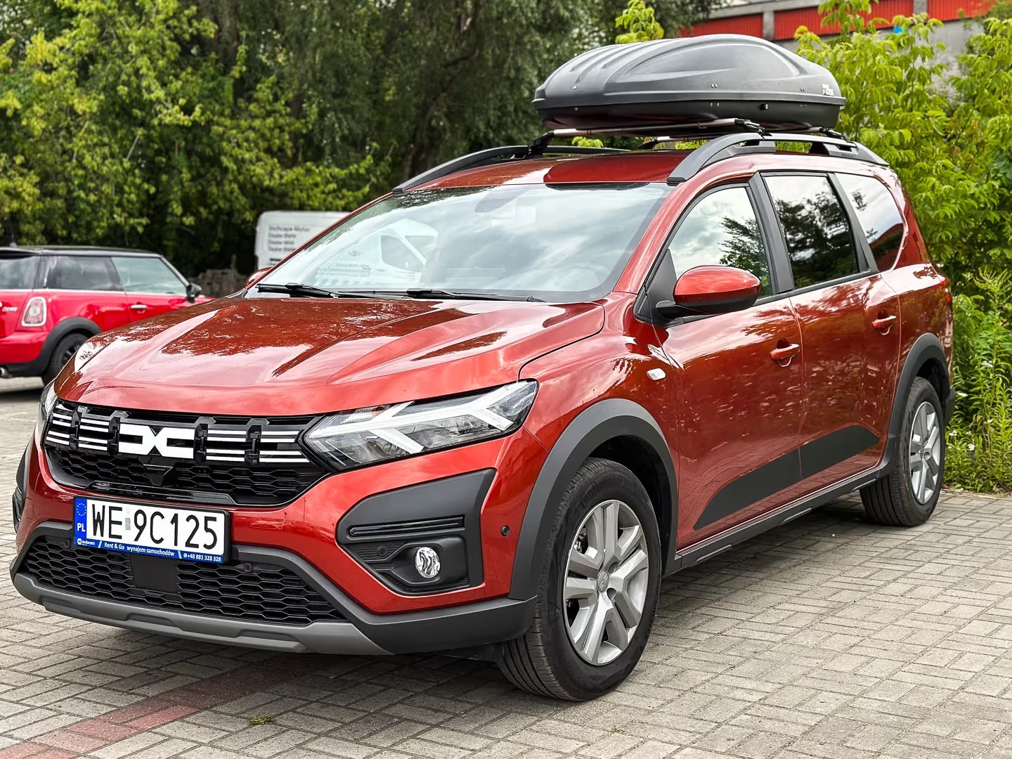 Czerwona Dacia Jogger z bagażnikiem dachowym na kostce brukowej, z widoczną tablicą rejestracyjną i fragmentem zieleni w tle. Auto 7-osobowe, idealne na wynajem.