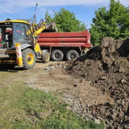 Żółta koparko-ładowarka i czerwona wywrotka podczas prac ziemnych na trawiastym terenie. W tle widoczne drzewa i słupy energetyczne.