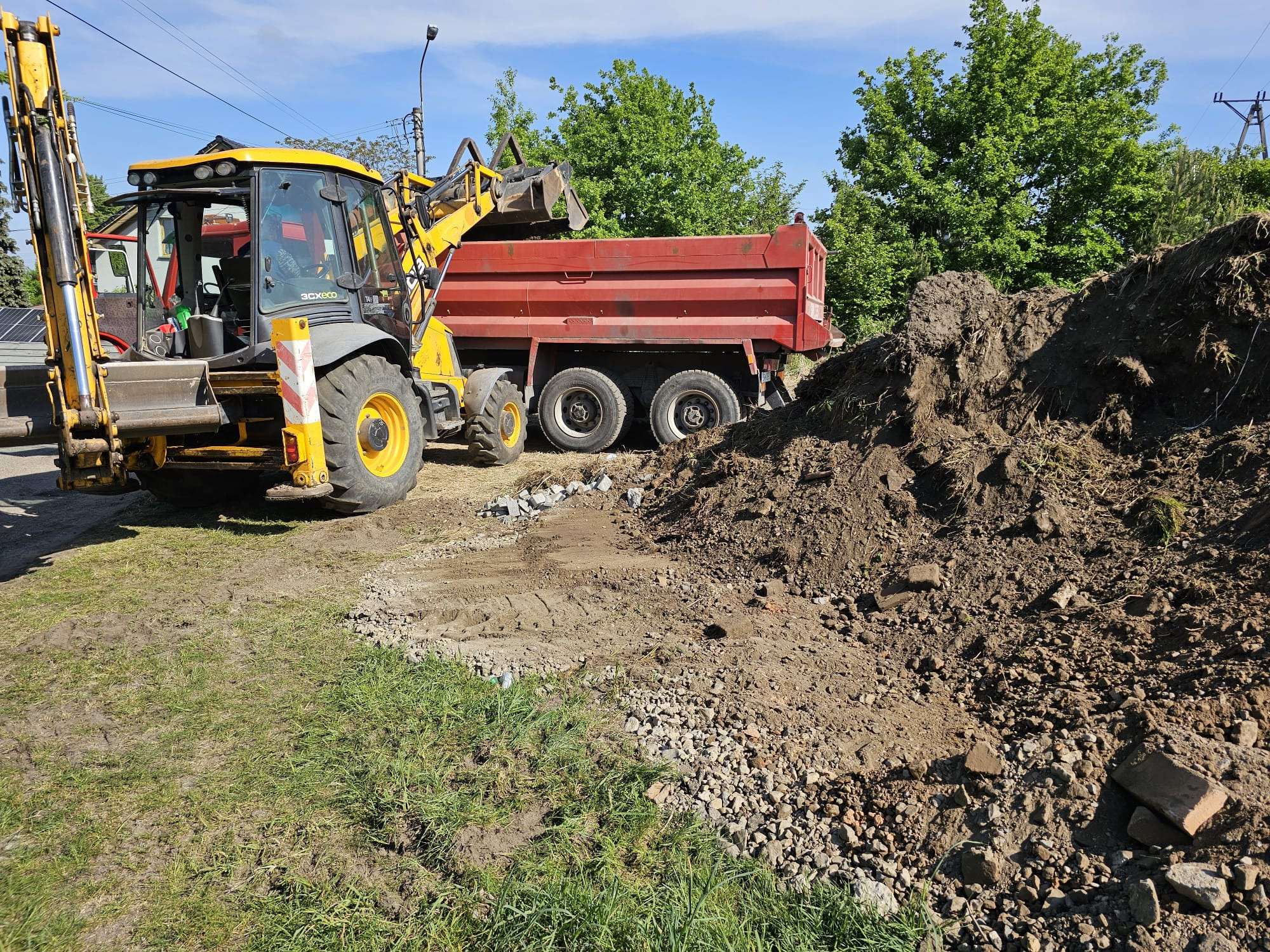 Żółta koparko-ładowarka i czerwona wywrotka podczas prac ziemnych na trawiastym terenie. W tle widoczne drzewa i słupy energetyczne.