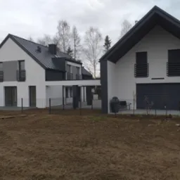 Nowoczesny, parterowy dom z białymi ścianami i grafitowym dachem blachodachówką. Dwa budynki połączone zadaszeniem. Surowy teren przed domem.