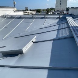 DS Dachbud - Daniel Sosiński - Błyszczący, szary dach z blachy trapezowej z metalową kładką serwisową. Widoczne elementy wentylacyjne i systemy odwadniające. W tle zabudowa miejska i błękitne niebo.