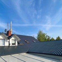 DS Dachbud - Daniel Sosiński - Ciemny, blaszany dach z lukarnami i kominem na tle błękitnego nieba. Widoczne rynny i antena satelitarna. Dach po renowacji, z nowym pokryciem.