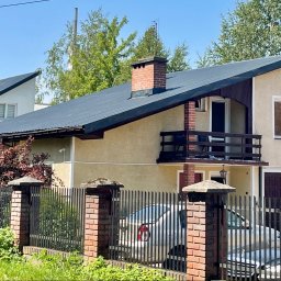 DS Dachbud - Daniel Sosiński - Dwupiętrowy dom z ciemnym dachem i ceglanym kominem, widziany zza czarnego, metalowego ogrodzenia z ceglanymi słupkami. Widoczne drzewa i zaparkowane samochody.