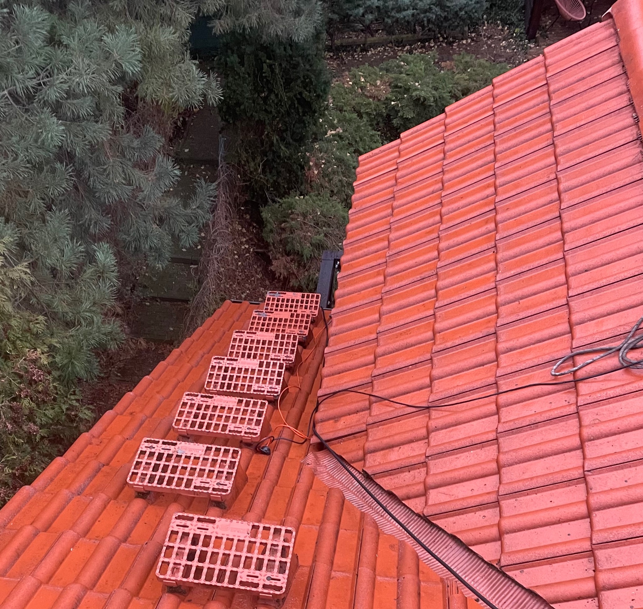 Czerwony dach z dachówki ceramicznej z zamontowanymi stopniami kominiarskimi, widok z góry, obok zieleń drzew. Elementy instalacji na dachu.