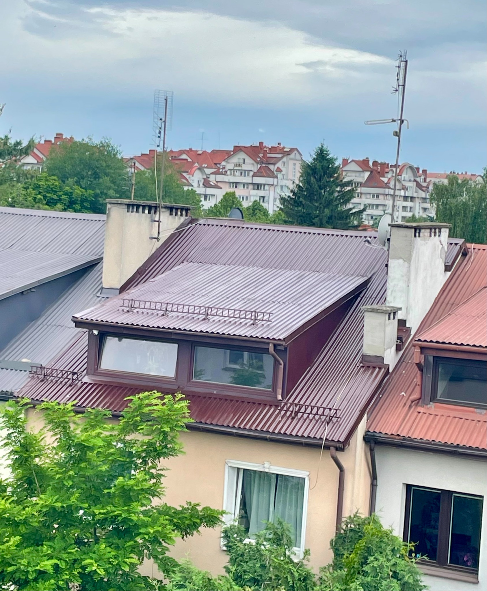 Czerwono-brązowy dach z blachy falistej, okno dachowe z barierką, komin i antena na tle zabudowy miejskiej i zieleni. Widok z góry.