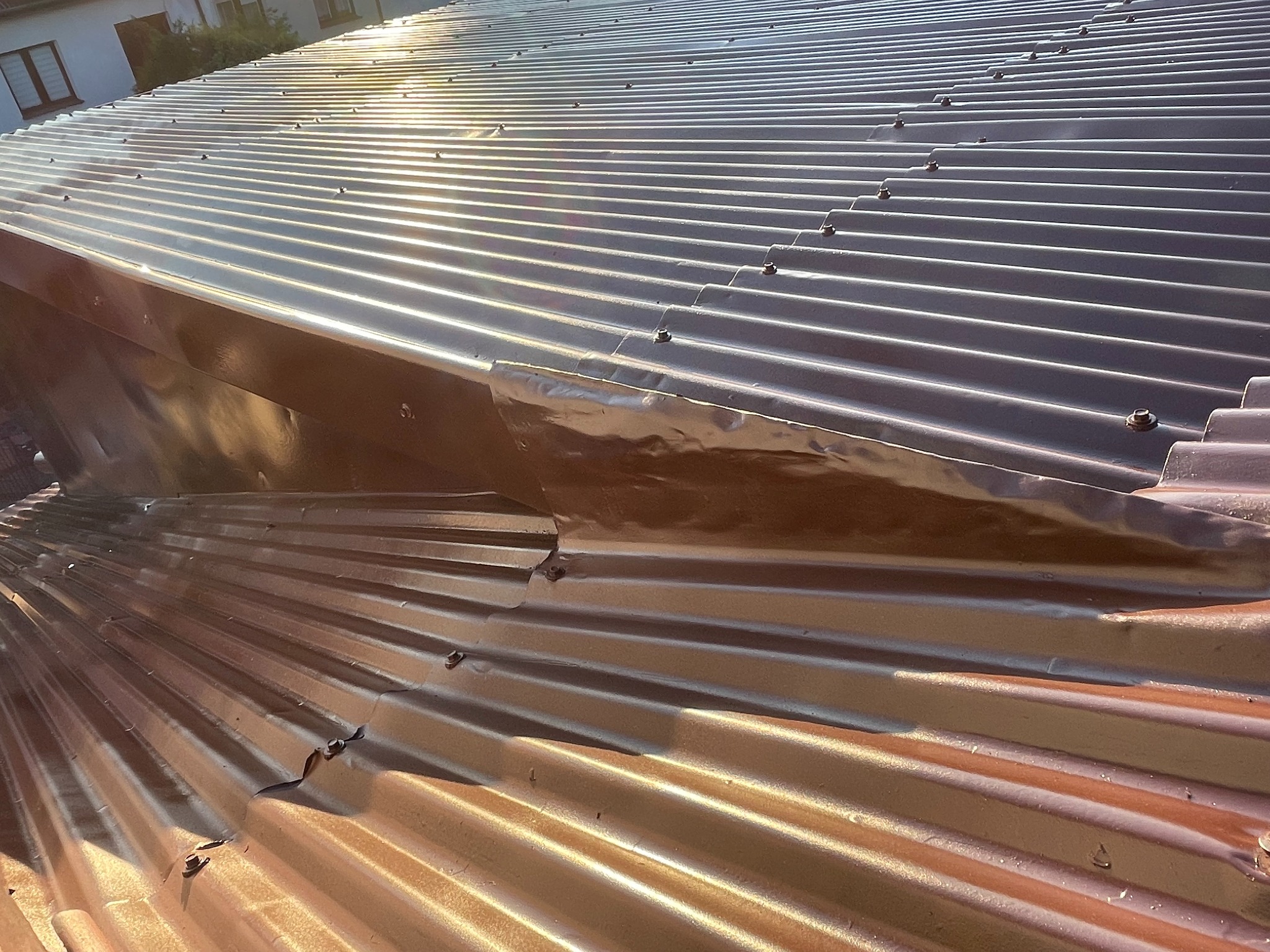 Błyszczący, brązowo-srebrny dach z blachy trapezowej, z widocznymi śrubami mocującymi i fragmentem budynku w tle, oświetlony promieniami słonecznymi.