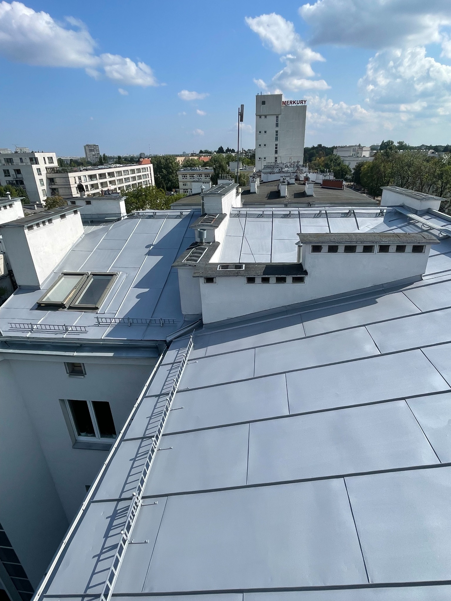 Błyszczący, metalowy dach budynku z lukarnami i kominami, widoczny z góry w słoneczny dzień. Na dachu zamontowana drabina. W tle zabudowa miejska i budynek z napisem MERKURY.