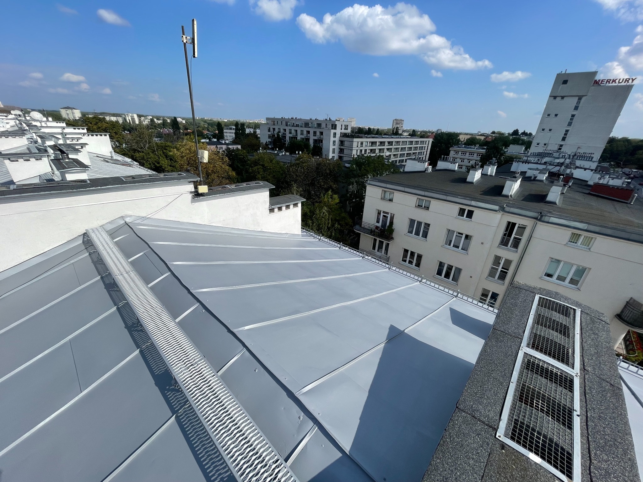 Szary, metalowy dach budynku z widoczną kratką przeciwśniegową i anteną na tle miejskiego krajobrazu w słoneczny dzień. Widok z góry.