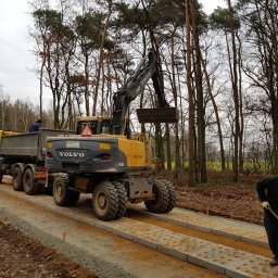 FERD SPÓŁKA Z OGRANICZONĄ ODPOWIEDZIALNOŚCIĄ - Żółto-szara koparka Volvo nabiera ziemię do wywrotki na tle lasu. Obok robotnik wyrównuje podłoże na drodze z betonowych płyt. Pochmurny dzień.