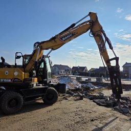 FERD SPÓŁKA Z OGRANICZONĄ ODPOWIEDZIALNOŚCIĄ - Żółta koparka SANY SY155W z hydraulicznym młotem pneumatycznym kruszy gruz na placu budowy. W tle zabudowa jednorodzinna i błękitne niebo.
