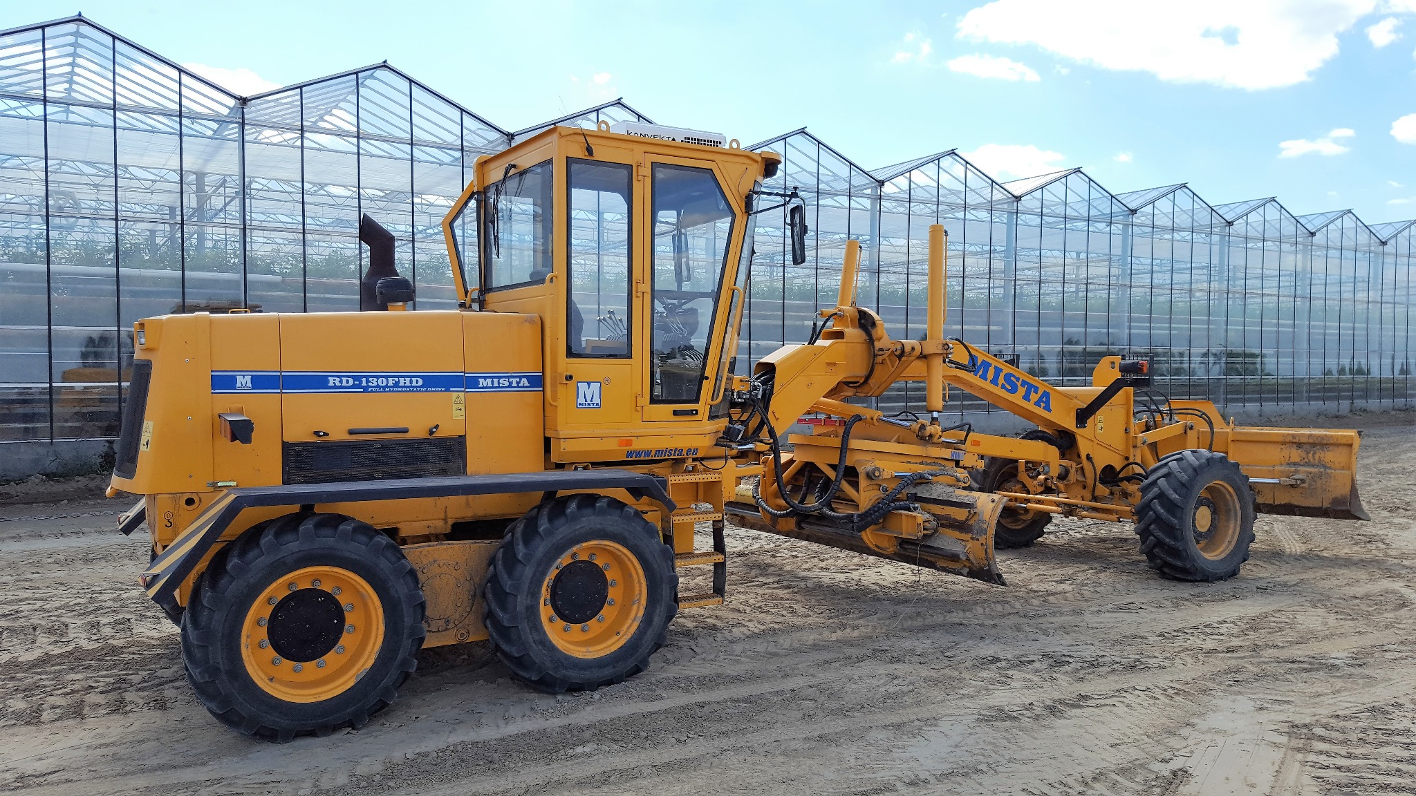 Żółty równiarka MISTA RD-130FHD na piaszczystym terenie, z szklarnią w tle. Maszyna gotowa do pracy, widoczne opony i lemiesz. Słoneczny dzień.
