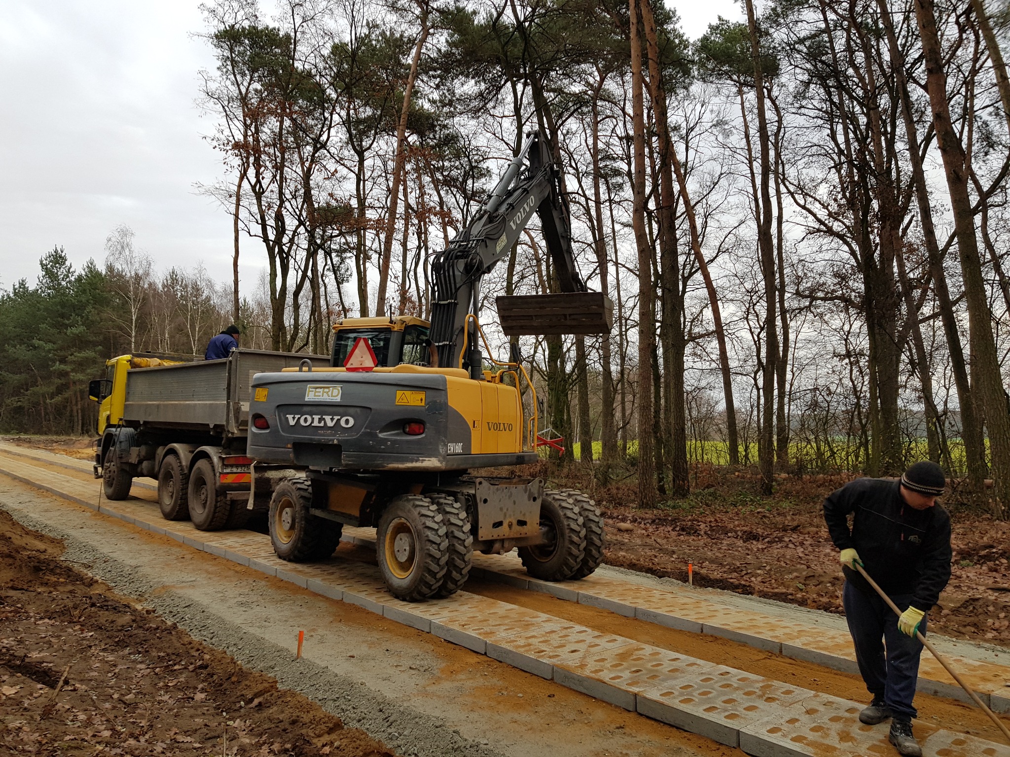 Żółto-szara koparka Volvo nabiera ziemię do wywrotki na tle lasu. Obok robotnik wyrównuje podłoże na drodze z betonowych płyt. Pochmurny dzień.