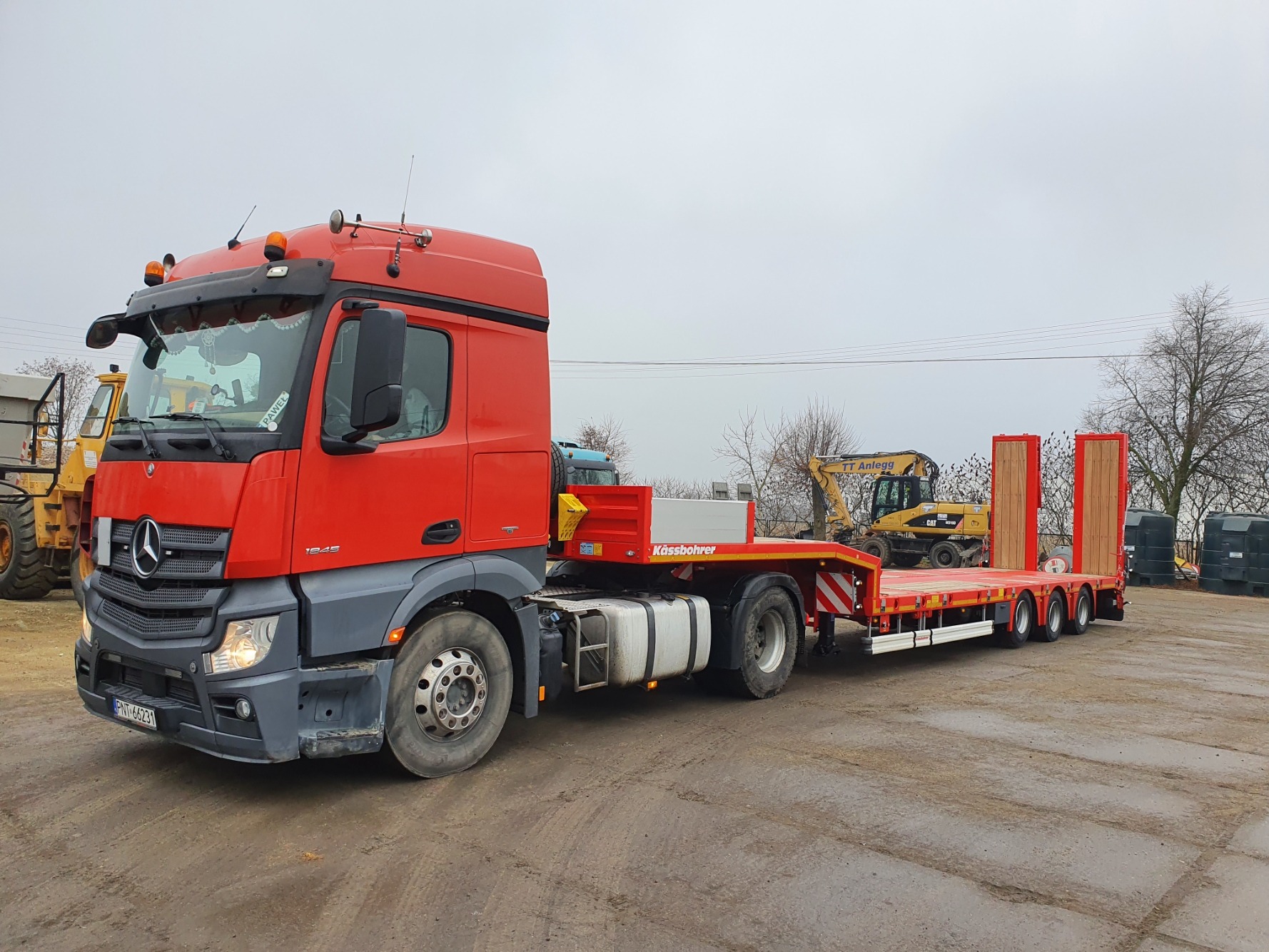 Czerwona ciężarówka z niskopodwoziową naczepą Kässbohrer, na której przewożona jest koparka. W tle widoczny krajobraz z drzewami i innymi maszynami budowlanymi.