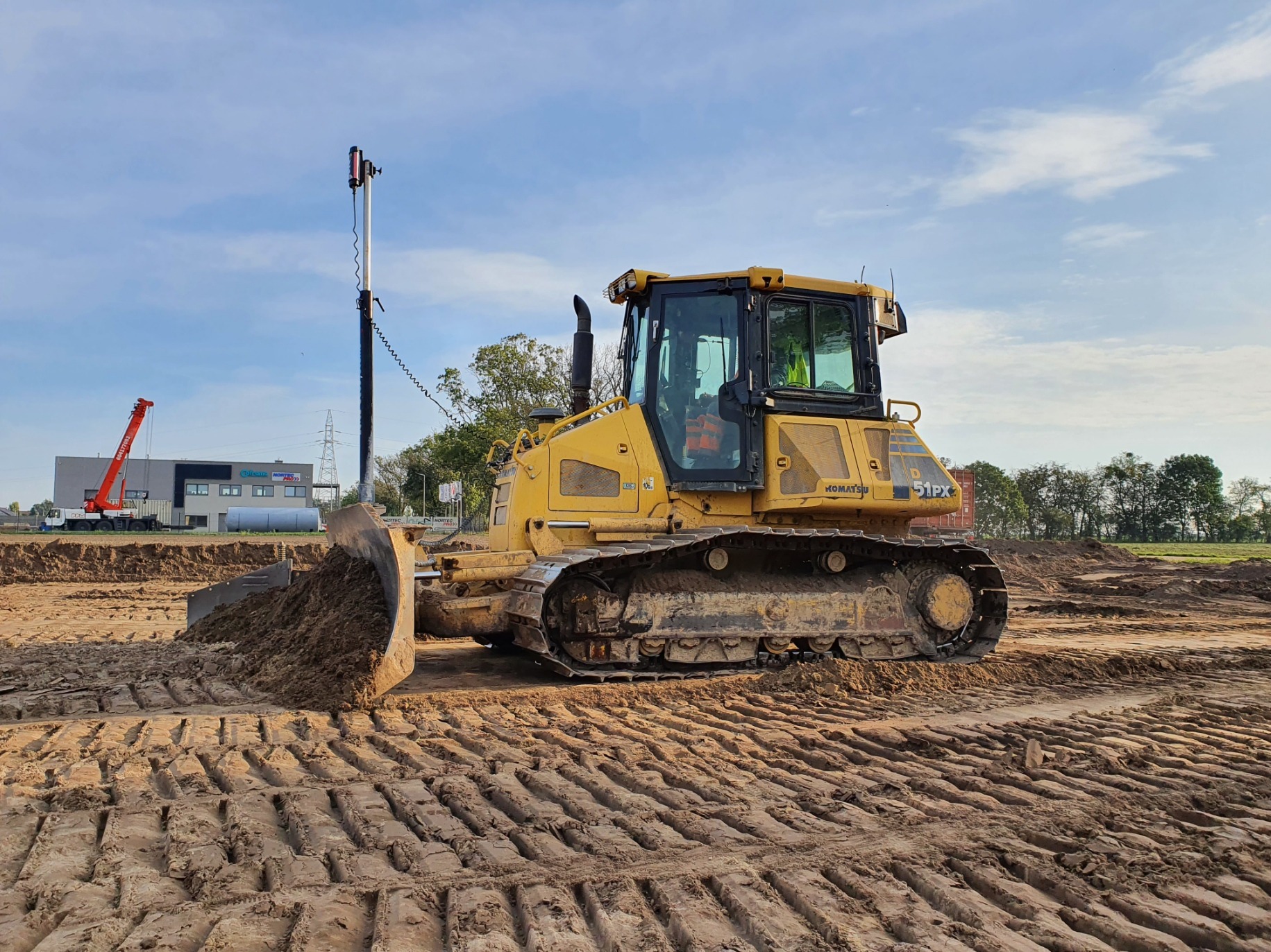 Żółty buldożer Komatsu 51PX z podniesioną łyżką na placu budowy. Widoczne ślady gąsienic na ziemi, w tle budynek i żuraw.