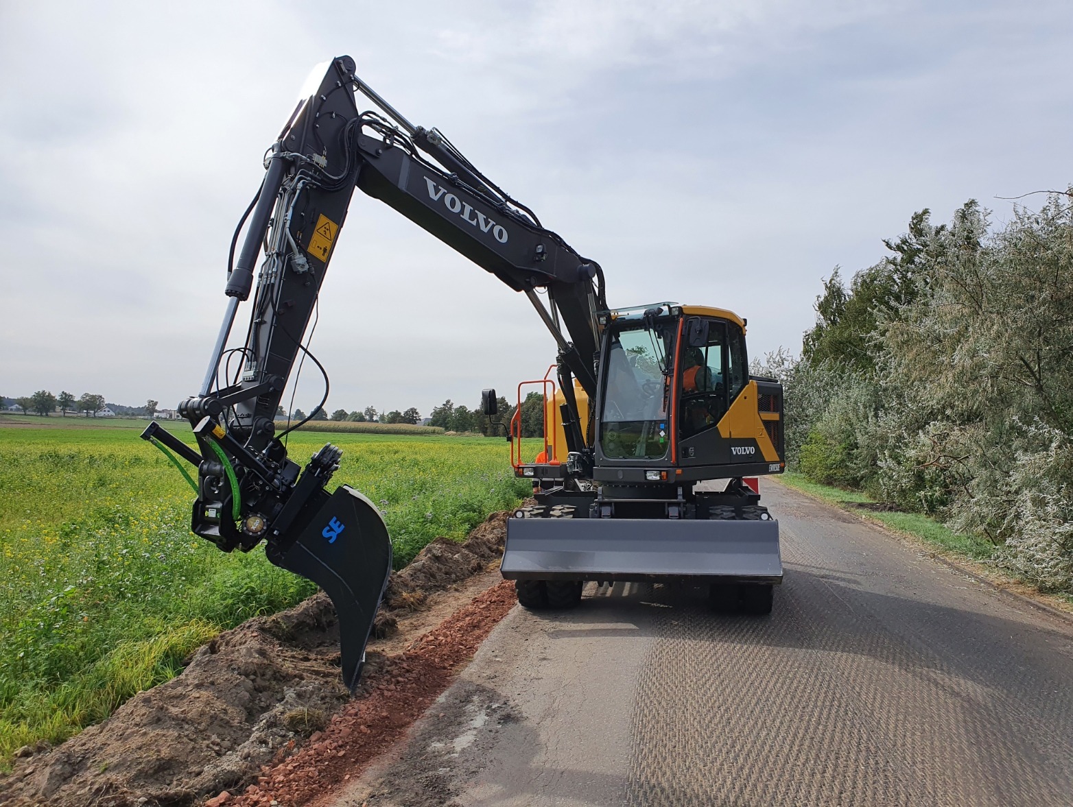 Żółto-czarna koparka Volvo na drodze z wykopanym rowem, zieleń w tle. Widoczna łyżka koparki tuż nad ziemią. Sprzęt gotowy do pracy.