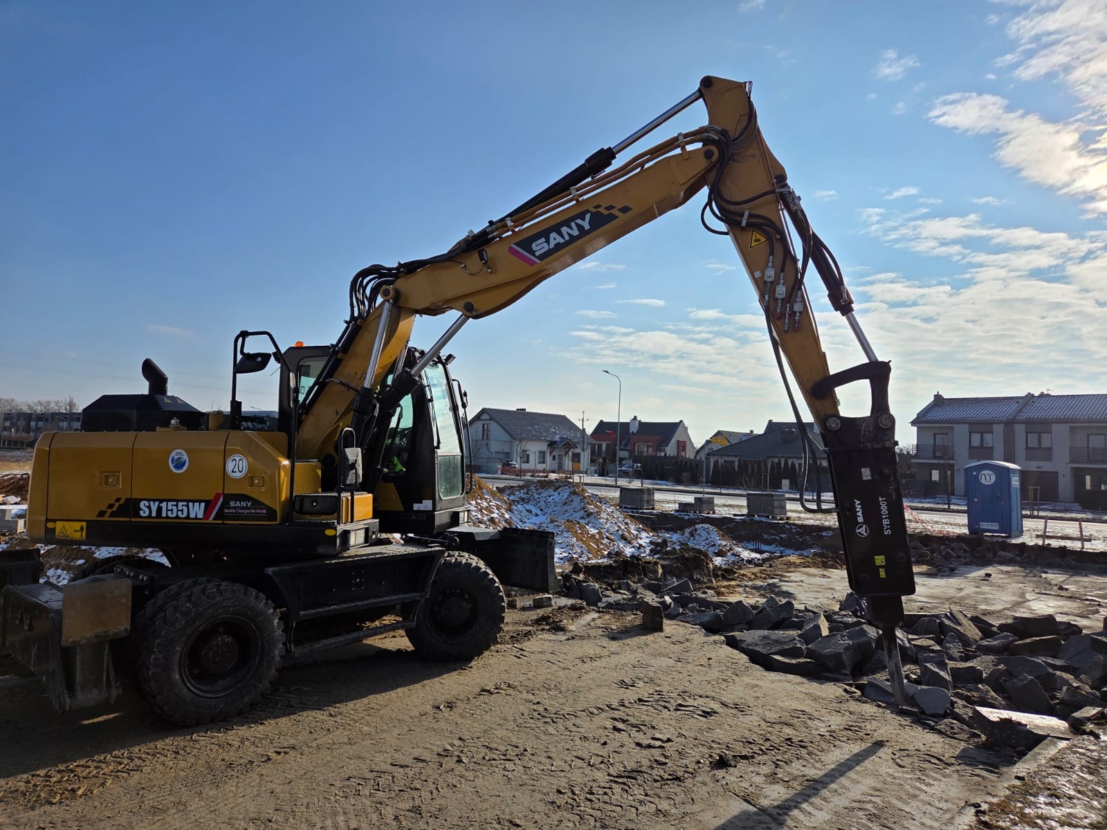Żółta koparka SANY SY155W z hydraulicznym młotem pneumatycznym kruszy gruz na placu budowy. W tle zabudowa jednorodzinna i błękitne niebo.