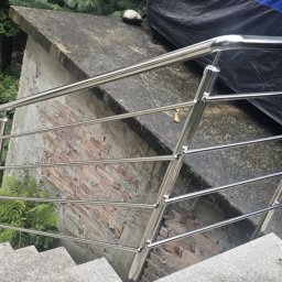 Metal-Bram Balustrady Nierdzewne ADAM URBAŚ - Polerowana balustrada ze stali nierdzewnej na schodach zewnętrznych, z widokiem na zieloną roślinność i fragment muru z cegły w tle.