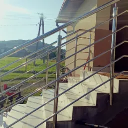 Zewnętrzne schody z jasnymi płytkami i lśniącą, metalową balustradą w słoneczny dzień. W tle dom z ogrodem i pagórkowaty krajobraz.