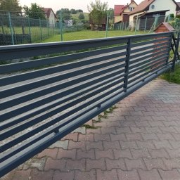 Metal-Bram Balustrady Nierdzewne ADAM URBAŚ - Nowoczesna, przesuwna brama posesyjna z poziomych, szarych profili. W tle zabudowa jednorodzinna i zieleń. Widoczne detale konstrukcyjne i wykończenie.