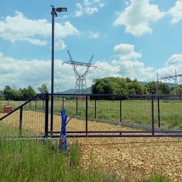Metal-Bram Balustrady Nierdzewne ADAM URBAŚ - Metalowa brama wjazdowa z siatki na posesję, z widoczną infrastrukturą energetyczną w tle. Teren wysypany kamieniami.